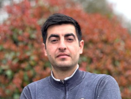 KI generiert: Ein Mann in grauem Pullover steht vor herbstlichem Hintergrund.