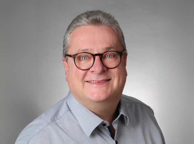 KI generiert: Ein Mann mit Brille lächelt in die Kamera, vor einem grauen Hintergrund.