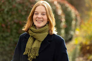 KI generiert: Eine lächelnde Person mit grünem Schal im Freien, unscharfer Hintergrund mit Büschen und Sonnenlicht.