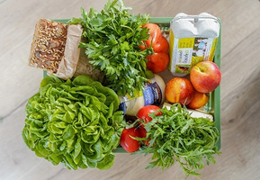 KI generiert: Ein Korb mit frischen Lebensmitteln: Brot, Kräuter, Salat, Tomaten, Eier, Äpfel.