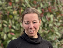 KI generiert: Eine Frau lächelt in die Kamera; der Hintergrund zeigt unscharfes Laub.