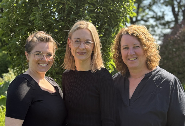 KI generiert: Drei Personen lächeln im Freien vor einem Baum in die Kamera.
