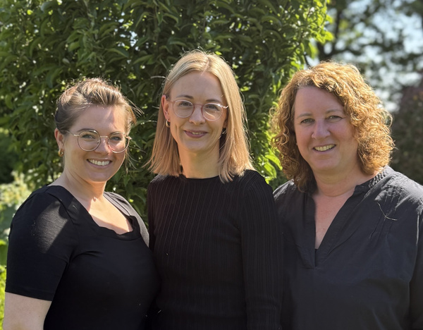 KI generiert: Drei Frauen lächeln in die Kamera, stehen draußen vor einem Baum.