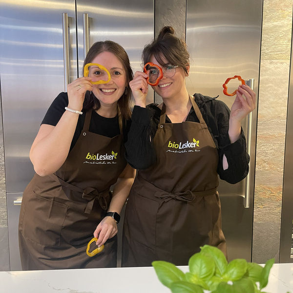 KI generiert: Zwei Personen in Schürzen posieren mit Paprikaringen vor ihren Augen. Text auf Schürzen: "bioLesker".