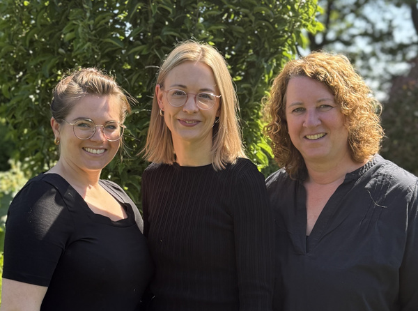 KI generiert: Drei lächelnde Personen stehen vor einem Baum im Freien.