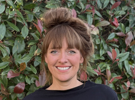 KI generiert: Eine lächelnde Person steht vor einer grünen Hecke mit rötlichen Blättern. Kein Text im Bild.